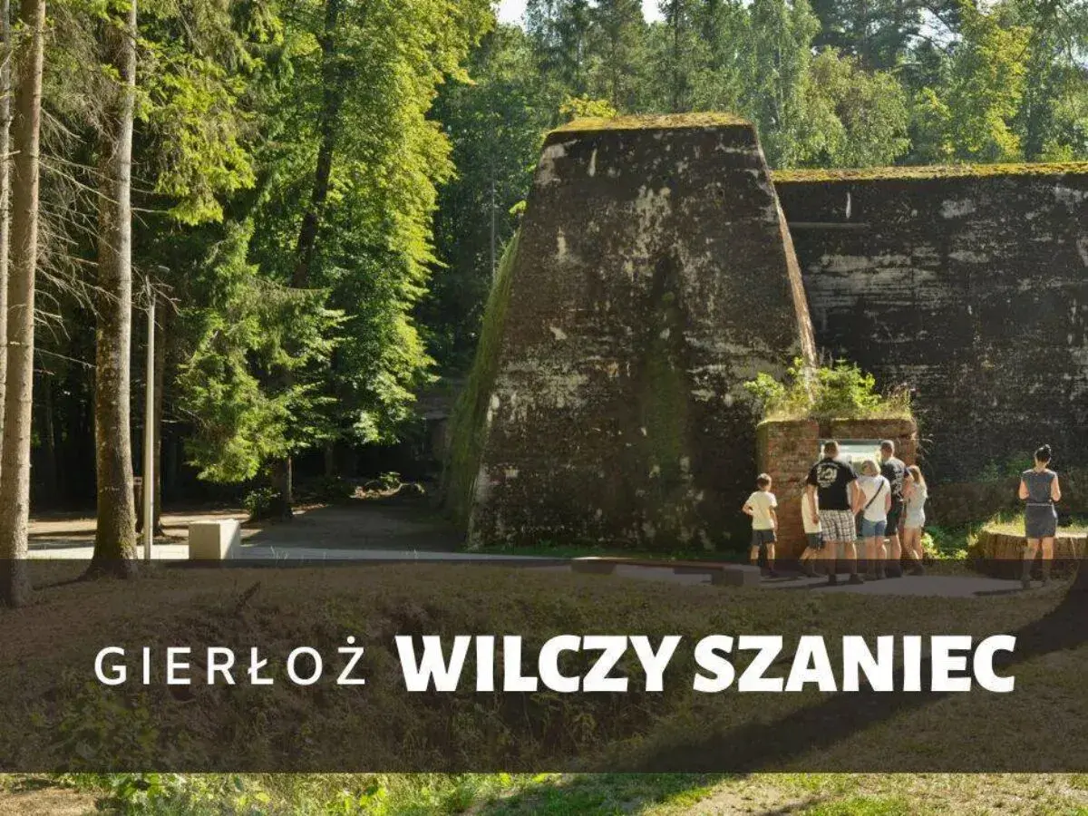 Grupa turystów zwiedza historyczny Wilczy Szaniec, otoczony bujną zielenią.