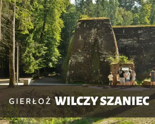 Grupa turystów zwiedza historyczny Wilczy Szaniec, otoczony bujną zielenią.