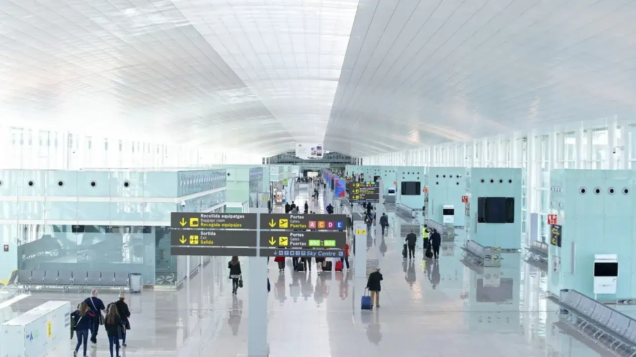 Jasny, przestronny terminal lotnisko Barcelona. Pasażerowie z bagażami przemieszczają się pod nowoczesnym, białym sufitem.