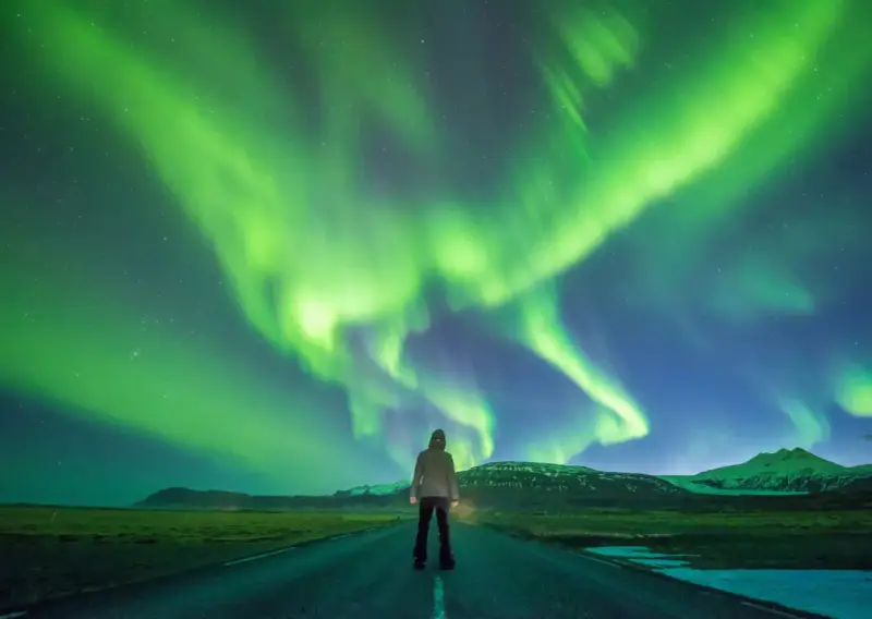 Człowiek stoi na drodze pod rozgwieżdżonym niebem, gdzie tańczy zorza polarna. Islandia, październik, pogoda idealna na takie widowisko.