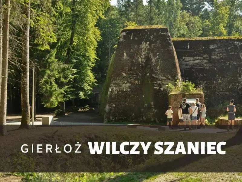 Grupa turystów zwiedza historyczny Wilczy Szaniec, otoczony bujną zielenią.